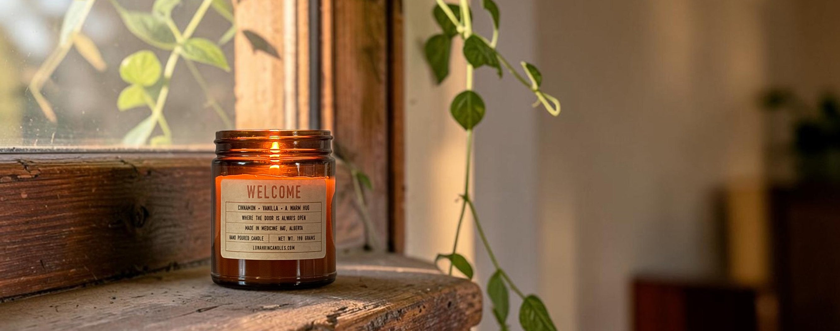 Lit Welcome candle on a wooden windowsill with a trailing green plant and warm afternoon light, Lunahrin Candles