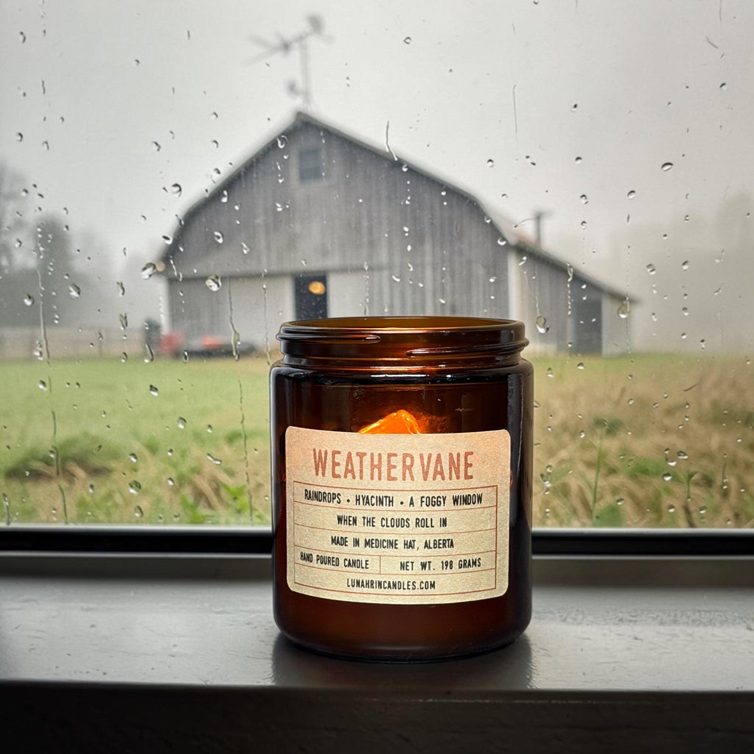 Rain-streaked window looking out at a barn with a weathervane on its roof, misty rain falling over an open field on an overcast day.